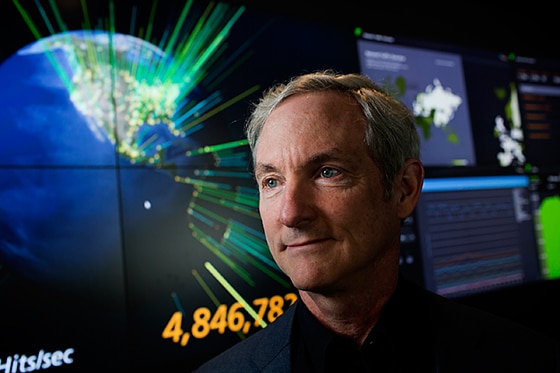 A man stands in front of a large data visualization display showing various metrics and global maps.