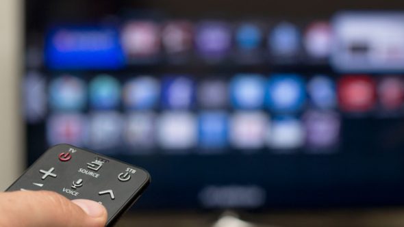 A hand holding a TV remote control in front of a television displaying various app icons.