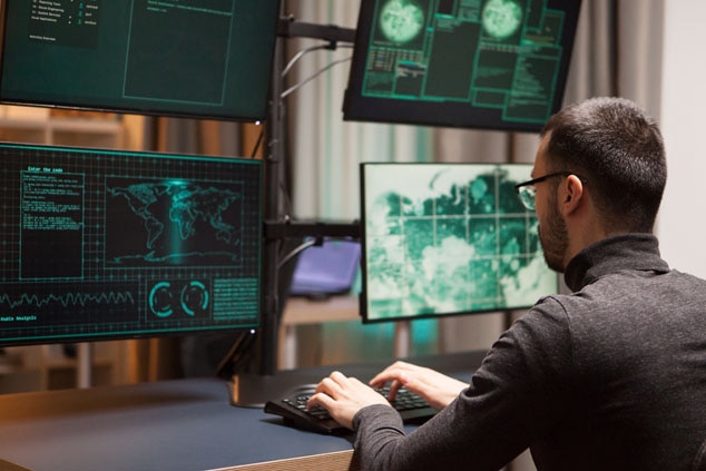 A cybersecurity analyst working at a desk with multiple screens displaying data and graphics.
