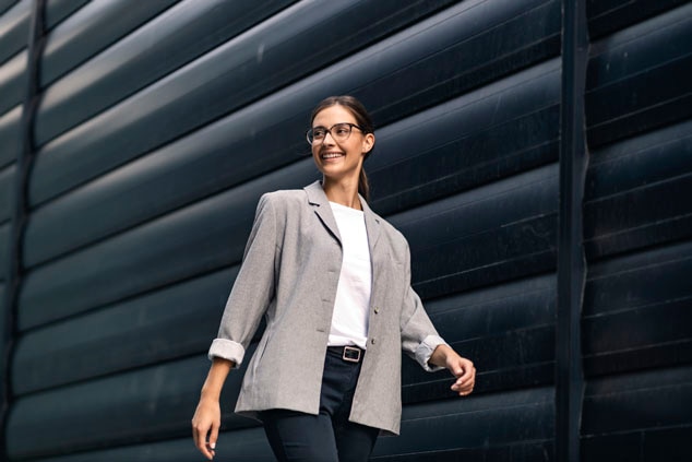 A woman in a gray blazer and white shirt,smiling while walking against a modern black wall.